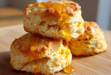 Three fluffy sweet potato biscuits stacked and drizzled generously with golden honey on a wooden surface.