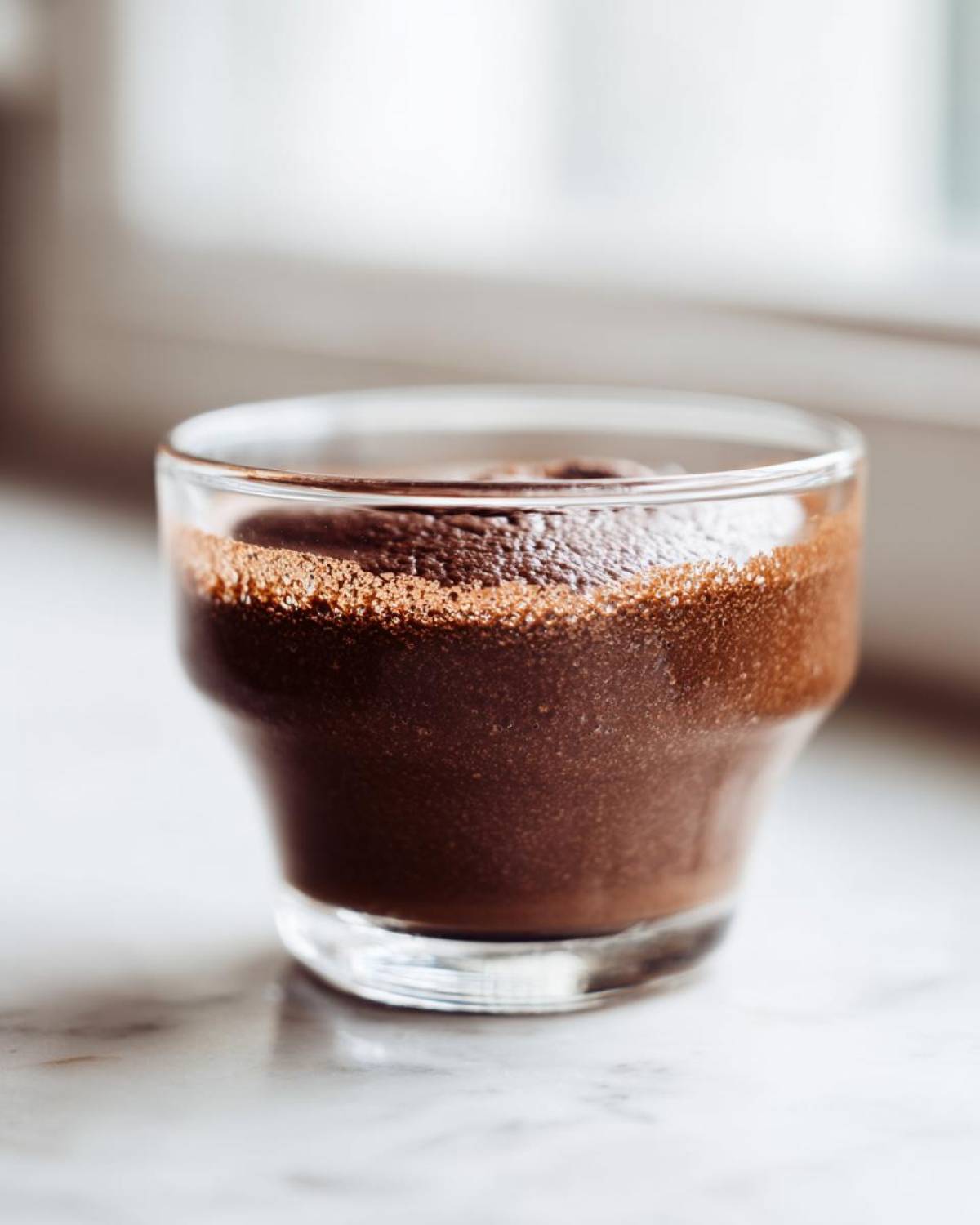 Close-up of rich, dark chocolate mousse served in a small clear glass on a white marble surface.