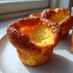 Close-up of a golden brown, perfectly risen yorkshire pudding with a crispy edge, sitting on a white plate.