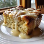 A thick slice of moist bread pudding topped generously with a sweet vanilla cream sauce, served on a white plate.