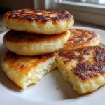 A stack of four golden brown, pan-fried irish potato farls on a white plate, one is broken open.