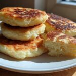 A stack of golden brown, pan-fried irish potato farls on a white plate, with one cut open showing the fluffy interior.