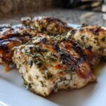 Four pieces of grilled chicken, glistening with herbs from the chicken marinade, resting on a white serving plate.