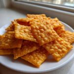 A pile of bright orange, square, textured homemade cheese crackers stacked on a small white plate near a window.