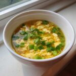 A white bowl filled with golden broth, wisps of cooked egg, and topped generously with chopped green onions, showcasing classic egg drop soup.