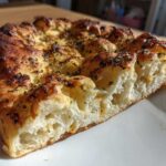 Close-up of a thick, airy slice of homemade pizza dough, baked golden brown and topped with dried herbs.