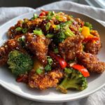 Close-up of crispy honey garlic chicken stir fry mixed with bright green broccoli and red/yellow bell peppers, topped with sesame seeds.