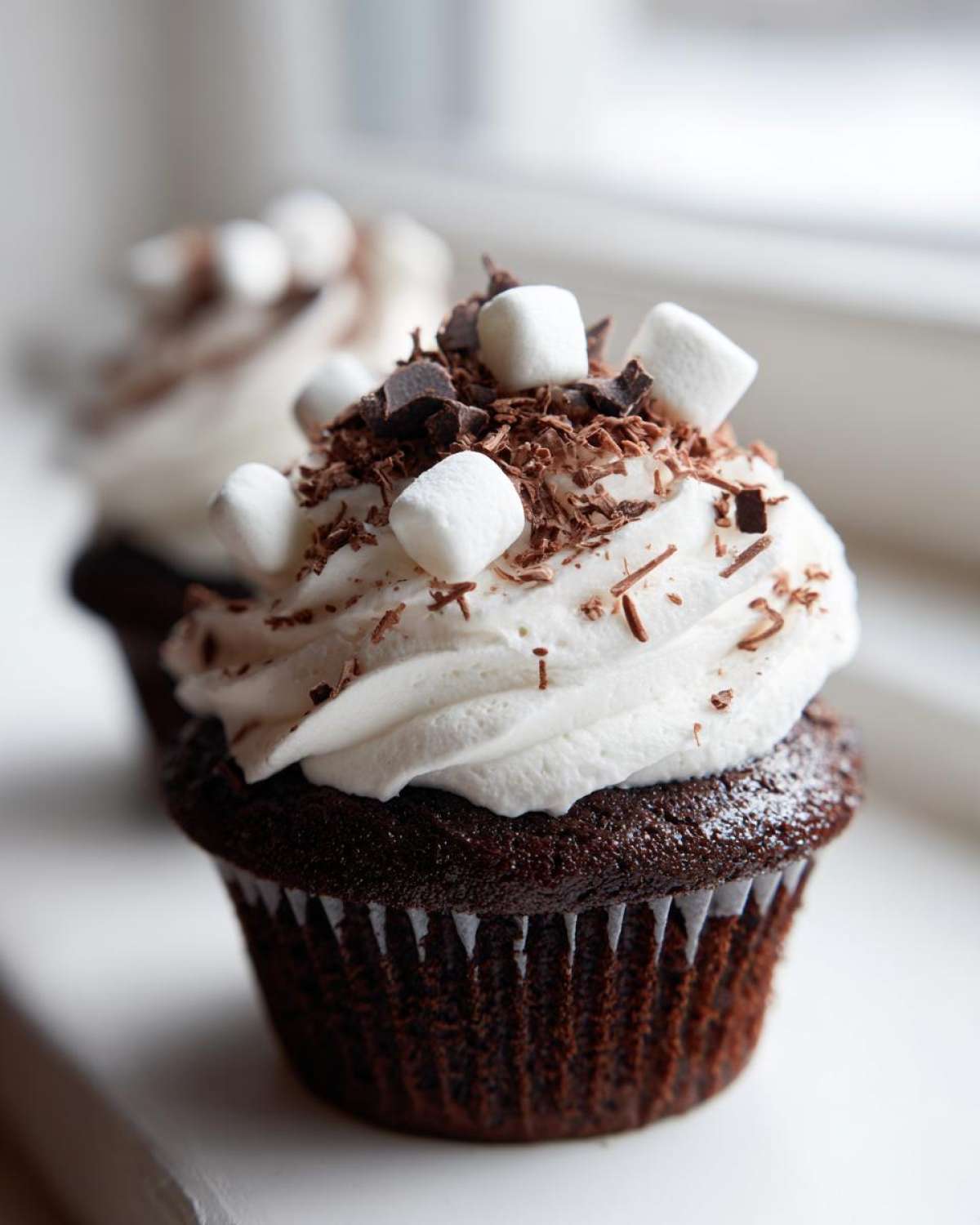 A close-up of a rich, dark chocolate hot chocolate cupcake topped with white frosting, mini marshmallows, and chocolate shavings.