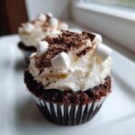 Close-up of a rich, dark chocolate hot chocolate cupcake topped with white frosting, mini marshmallows, and chocolate shavings.