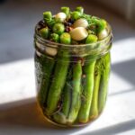 A glass jar tightly packed with bright green dilly beans, whole garlic cloves, dill sprigs, and spices, ready for pickling.