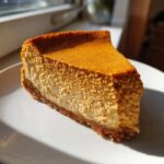 A close-up slice of rich, creamy keto pumpkin cheesecake with a graham cracker style crust, sitting on a white plate.