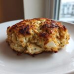 A single, golden-brown Maryland crab cake with visible lumps of crab meat, sitting on a white plate.