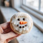 A hand holding one delicious melted snowman cookie decorated with white icing, chocolate chip eyes/mouth, and a candy carrot nose.