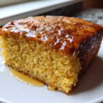 A close-up of a square slice of golden sourdough cornbread drizzled generously with honey or syrup on a white plate.