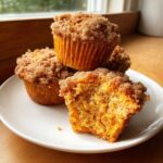 Close-up of moist sweet potato muffins stacked on a white plate, one is broken open showing the texture.