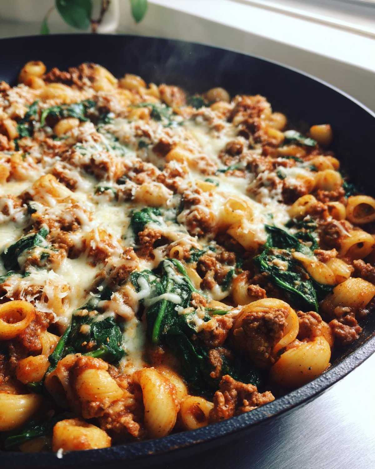 Close-up of steaming ground turkey pasta mixed with spinach and topped with melted cheese in a black skillet.