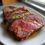 Close-up of three thick, medium-rare slices of prime rib seasoned with herbs and garlic on a white plate.