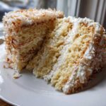 Close-up of a moist slice of coconut cake recipe layered with white frosting and covered in toasted coconut flakes.