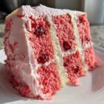 A moist slice of pink layered cherry cake with visible whole cherries and white/pink frosting on a white plate.