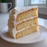 A tall, moist slice of three-layer vanilla cake recipe covered in thick white buttercream frosting.