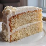 A close-up slice of moist vanilla cake recipe layered with creamy white frosting on a white plate.