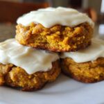 A stack of three soft pumpkin cookie treats topped generously with thick, white cream cheese frosting.