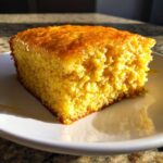 A close-up of a golden slice of moist sourdough cornbread dripping with honey on a white plate.