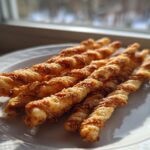 Close-up of several golden brown, twisted, spicy cheese straws resting on a white plate near a window.