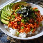 A close-up of a vibrant tuna rice bowl topped with spicy sauce, sliced avocado, nori, and green onions.