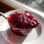 A spoonful of thick, glossy, deep red homemade raspberry jam held over a small jar.