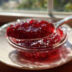 A spoonful of vibrant, glistening homemade raspberry jam held over a small glass bowl, catching sunlight.