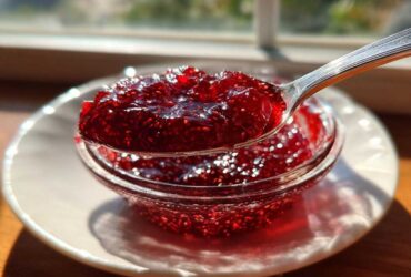 A spoonful of vibrant, glistening homemade raspberry jam held over a small glass bowl, catching sunlight.