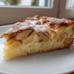 A close-up of a moist slice of Swedish apple cake, featuring tender apple pieces baked into a light sponge, dusted with powdered sugar.