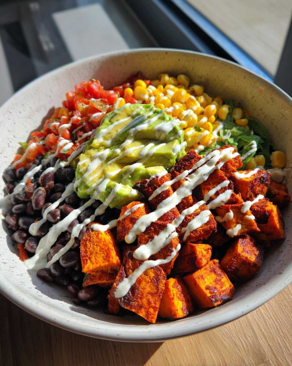 A vibrant sweet potato taco bowl featuring seasoned sweet potato cubes, black beans, corn, pico de gallo, avocado, and a white cream drizzle.