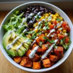 A vibrant sweet potato taco bowl featuring roasted sweet potato cubes, black beans, corn salsa, avocado slices, and a drizzle of white sauce.