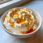 A white bowl filled with creamy whipped ricotta dip topped with drizzled hot honey and fresh rosemary.
