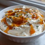 A white bowl filled with creamy whipped ricotta dip, topped with a generous drizzle of hot honey and fresh herbs.