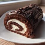 A cross-section slice of a rich chocolate yule log cake showing the dark sponge and white cream swirl filling.