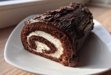 A cross-section slice of a rich chocolate yule log cake showing the dark sponge and white cream swirl filling.