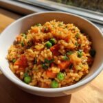 A close-up of vibrant, orange-colored spanish rice mixed with bright green peas and chopped carrots in a white bowl.