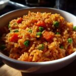 A close-up shot of fluffy, orange-tinted spanish rice mixed with bright green peas and diced carrots, served in a white bowl.