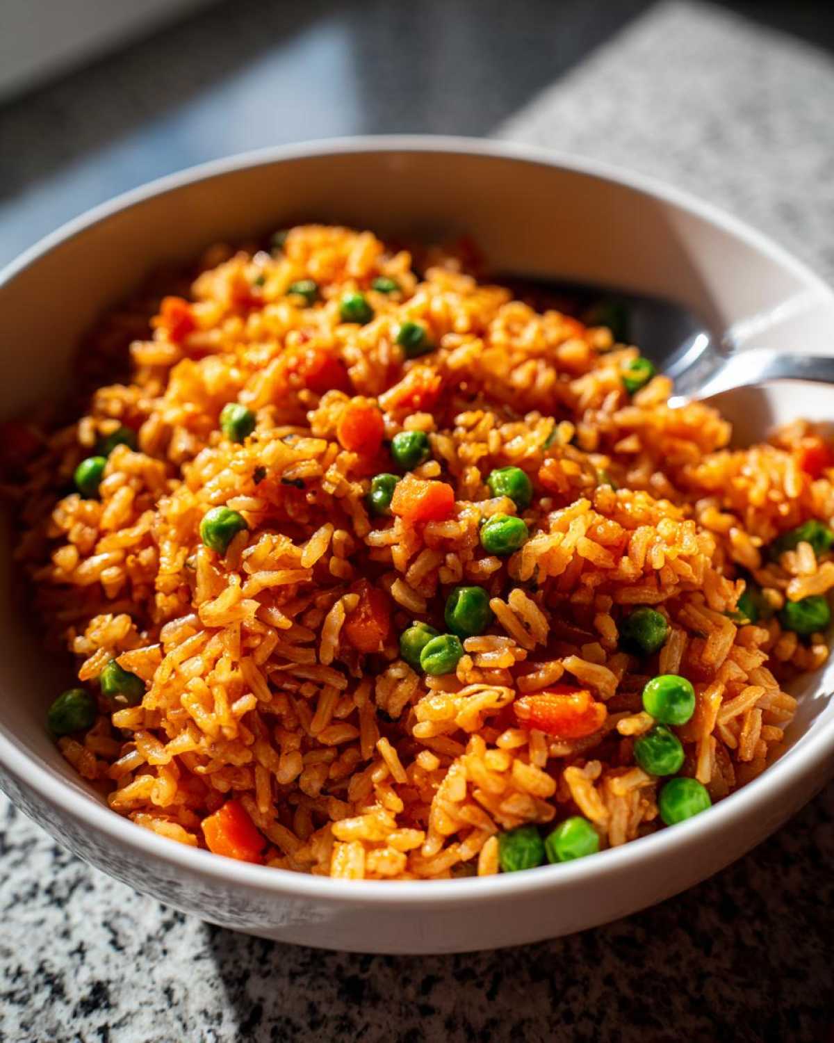 A close-up of bright orange spanish rice mixed with green peas and diced carrots in a white bowl.