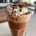 Close-up of a tall glass filled with frothy frozen hot chocolate topped with whipped cream and chocolate shavings.