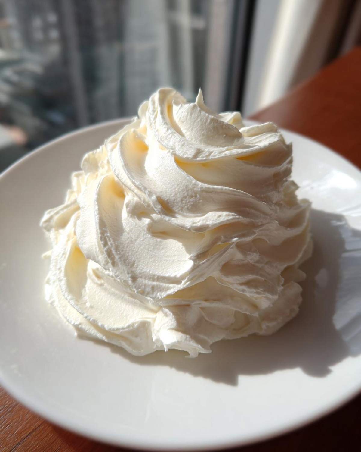 A beautiful swirl of thick, white cream cheese frosting piped onto a white plate, highlighted by bright sunlight.