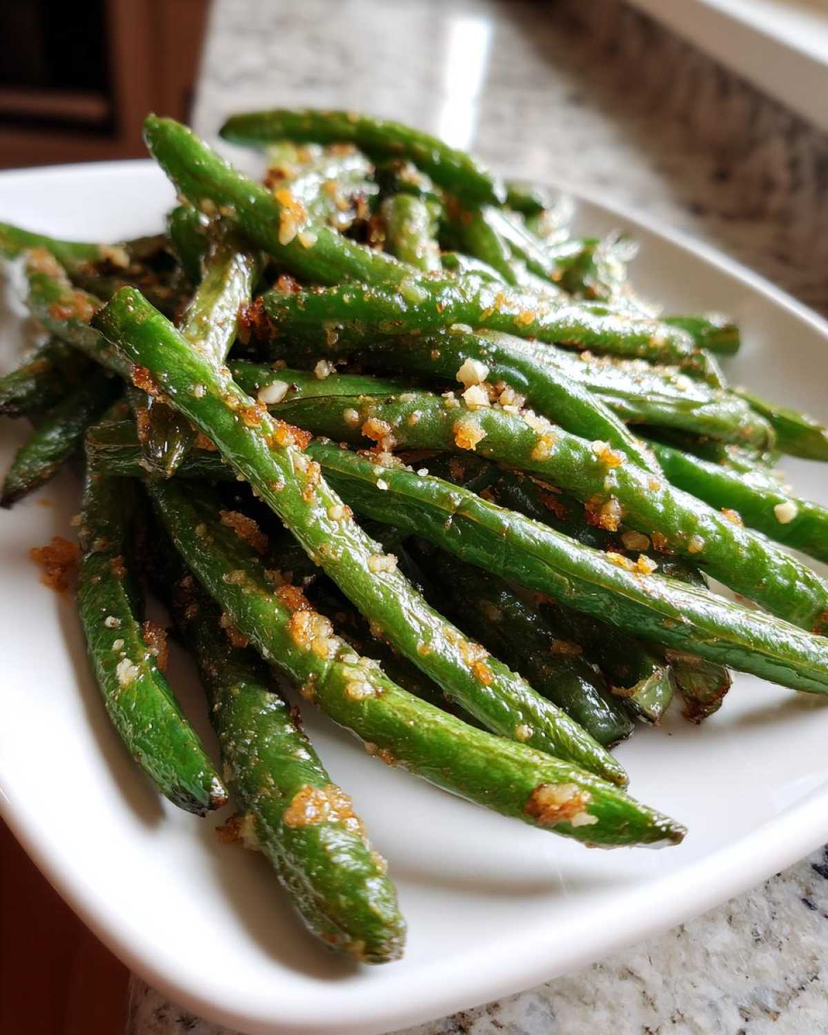 Close-up of bright green beans tossed with toasted garlic and seasonings on a white serving dish.