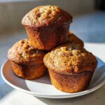 A stack of four perfectly baked, golden brown banana nut muffins sitting on a white plate.