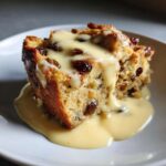 Close-up of a square slice of raisin bread pudding generously topped with creamy vanilla custard sauce.