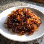 A close-up of deeply browned, glossy caramelized onions piled high on a small white dish.