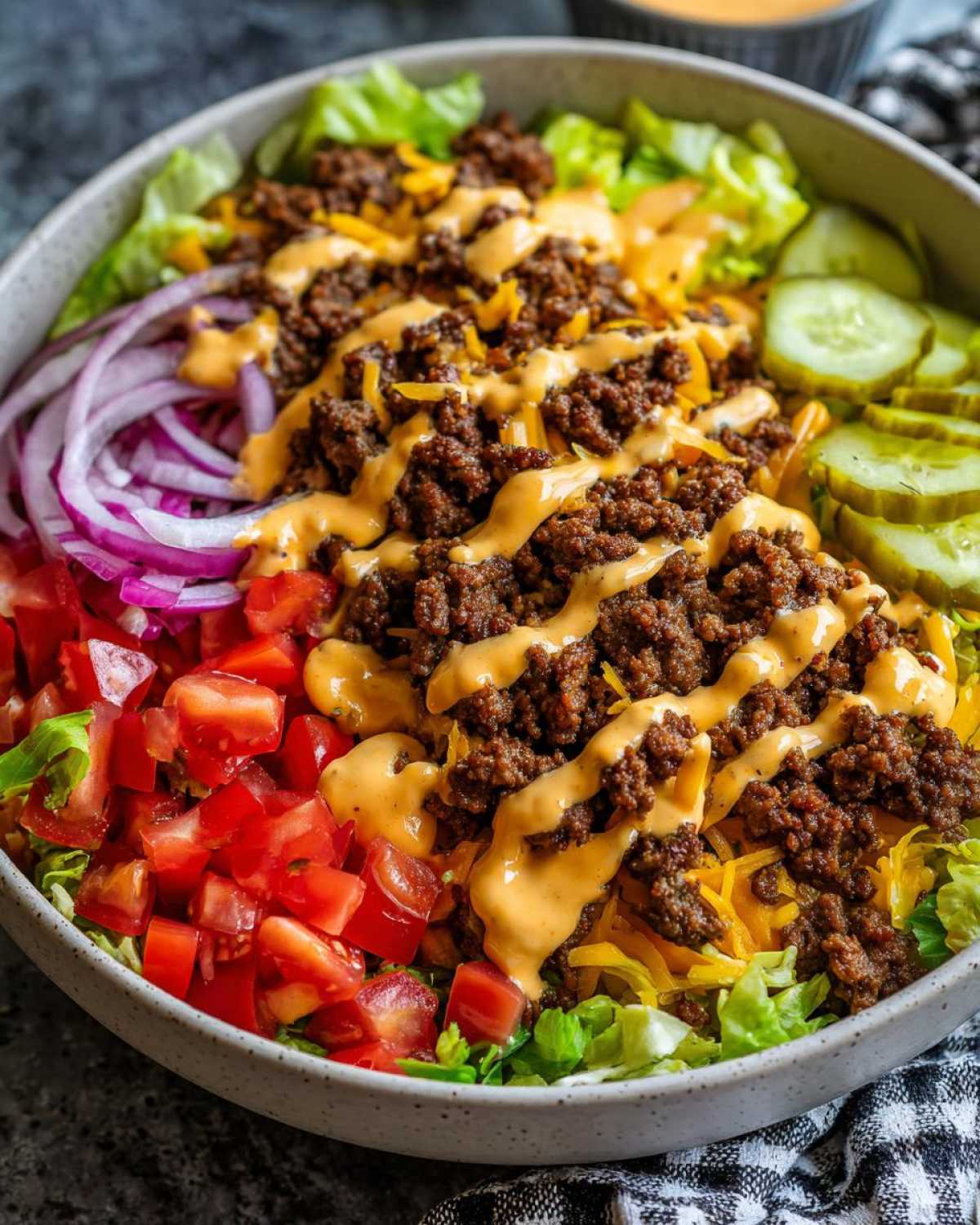 A vibrant cheeseburger bowl featuring seasoned ground beef, lettuce, tomatoes, red onion, pickles, and a special sauce drizzle.