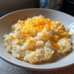 A close-up of a white bowl filled with creamy, amazing cheesy rice topped generously with melted shredded cheddar cheese.
